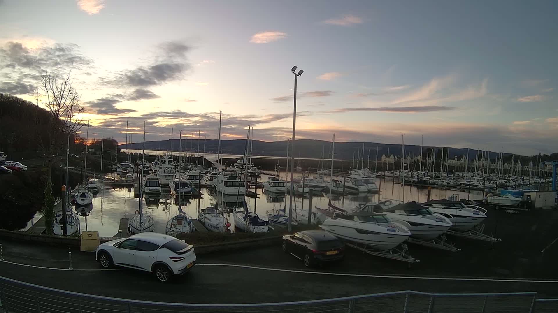 A marina filled with numerous boats of various sizes, is seen at sunset under a partly cloudy sky, with two cars parked nearby.