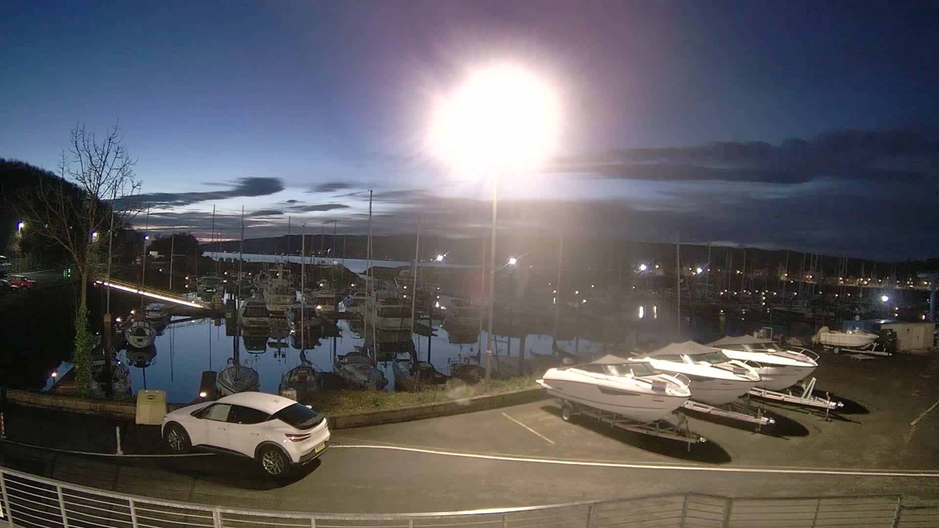 A white car is parked near several boats in a marina at twilight under a mostly clear sky.