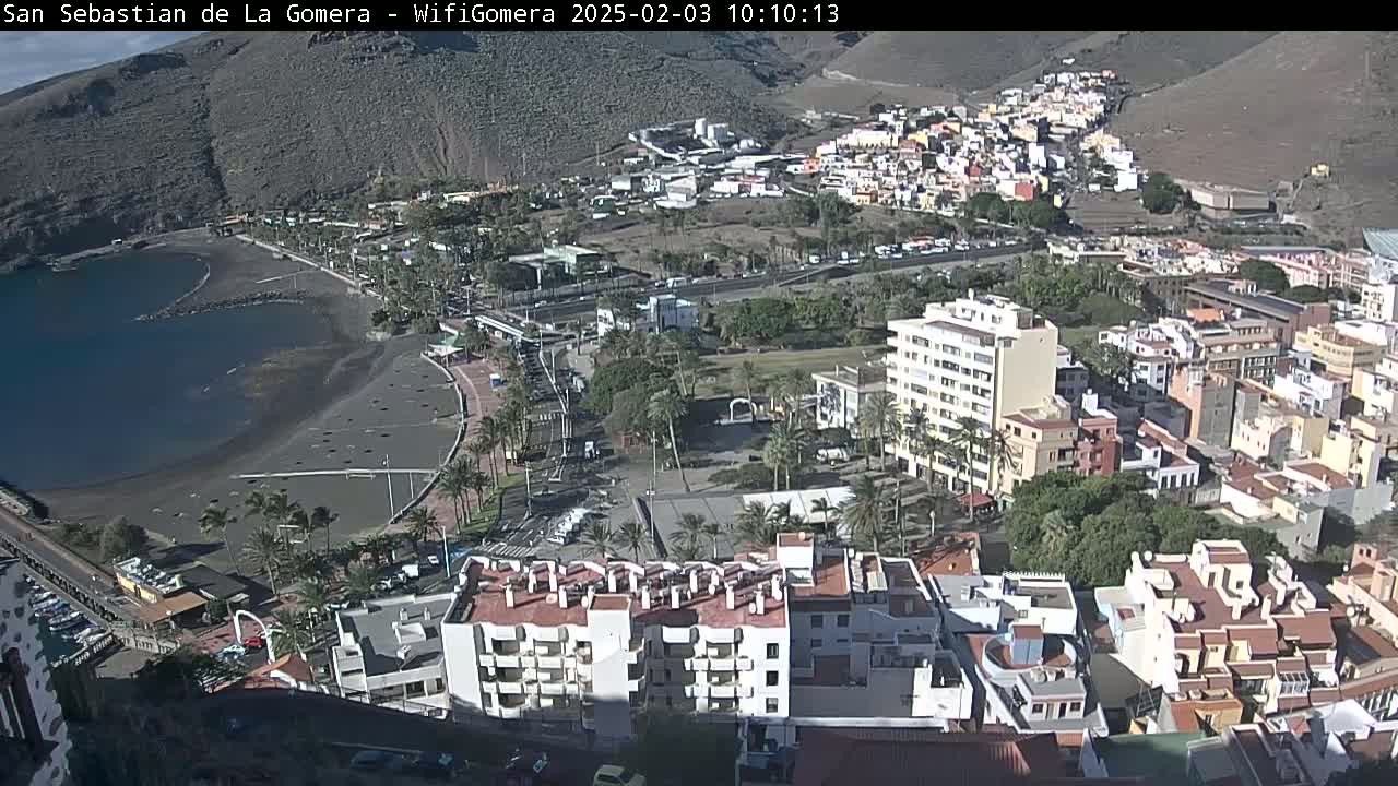 A coastal town with a dark-sand beach and calm waters nestled against a mountainous backdrop on a sunny day.