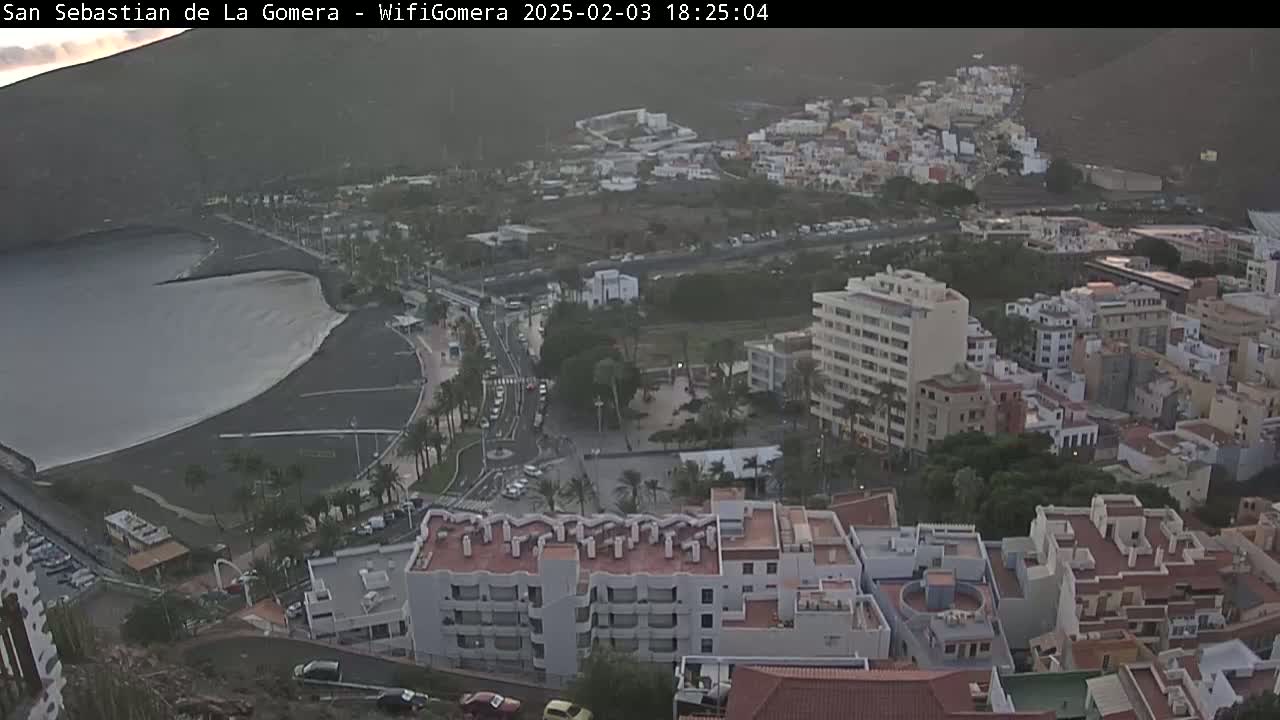 An aerial view of a coastal town at dusk, showing a dark-grey ocean, a sandy beach, roads with cars, and numerous buildings rising up a hillside.