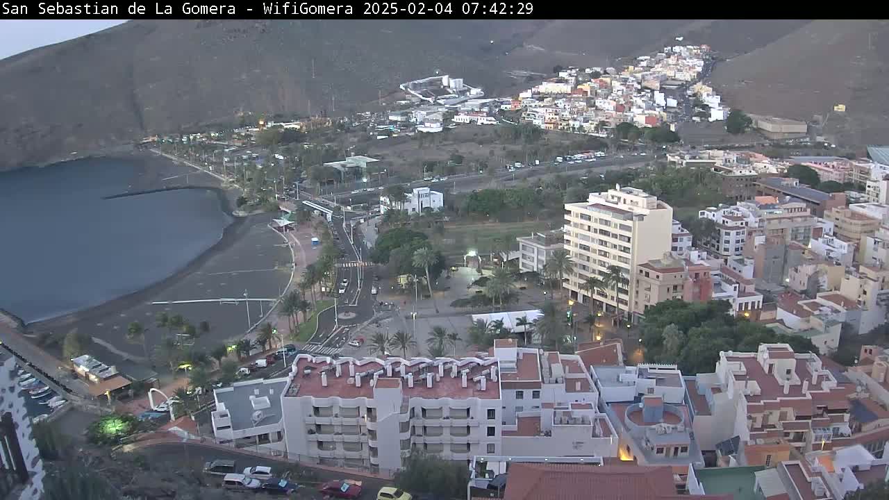 An aerial view of a coastal town at dawn shows a dark-colored beach next to a calm body of water, with buildings and roads extending uphill toward a mountainside.