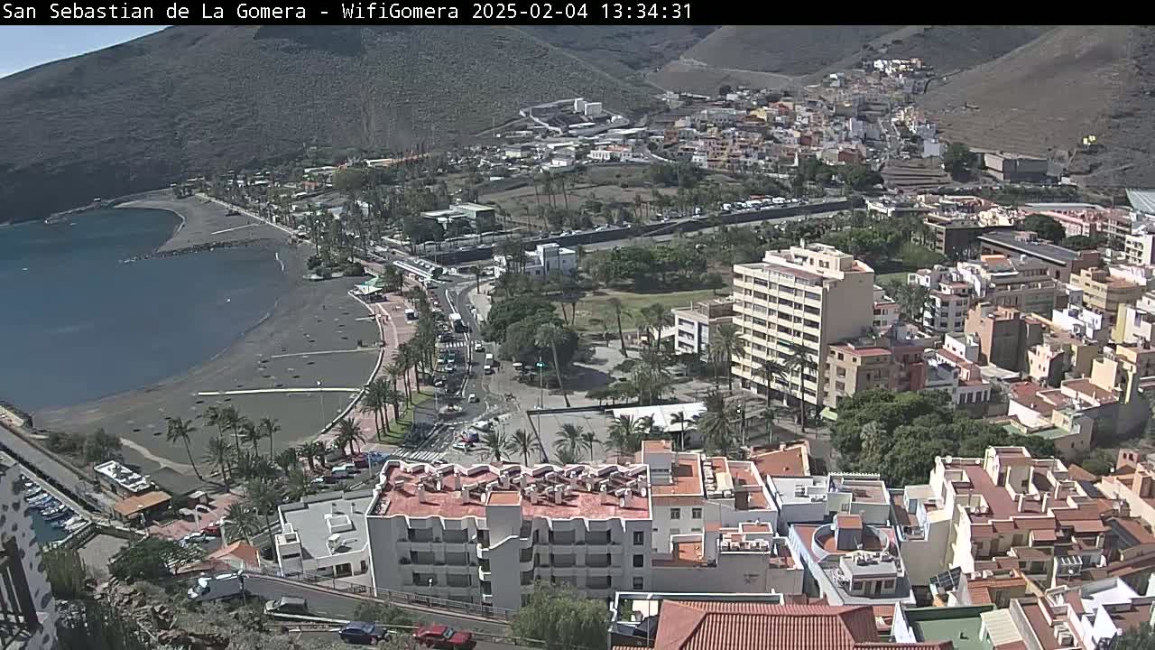 An aerial view shows a coastal town on a sunny day, with a dark-sand beach next to a calm bay and many buildings extending up a hillside.