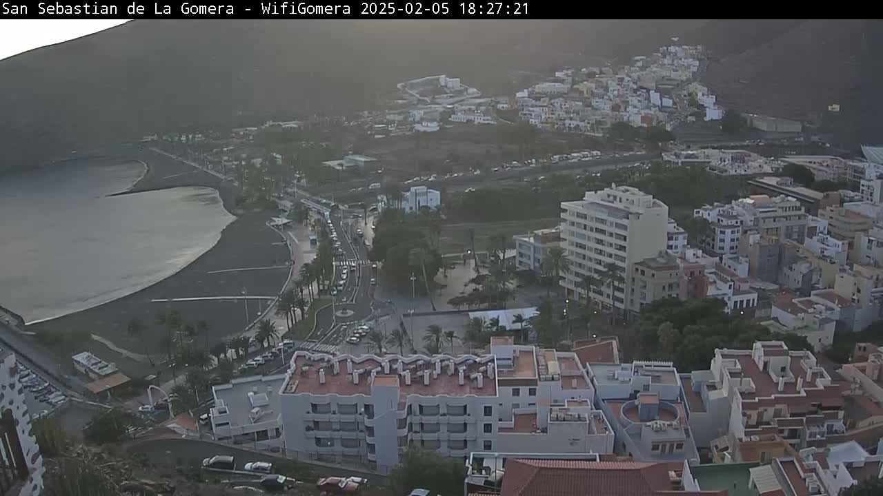 A hazy, dusk-lit coastal town with a dark-sand beach, several multi-story buildings, and roads filled with cars.