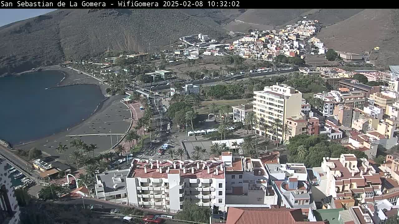 A coastal town with a black sand beach and numerous buildings is seen on a sunny day.