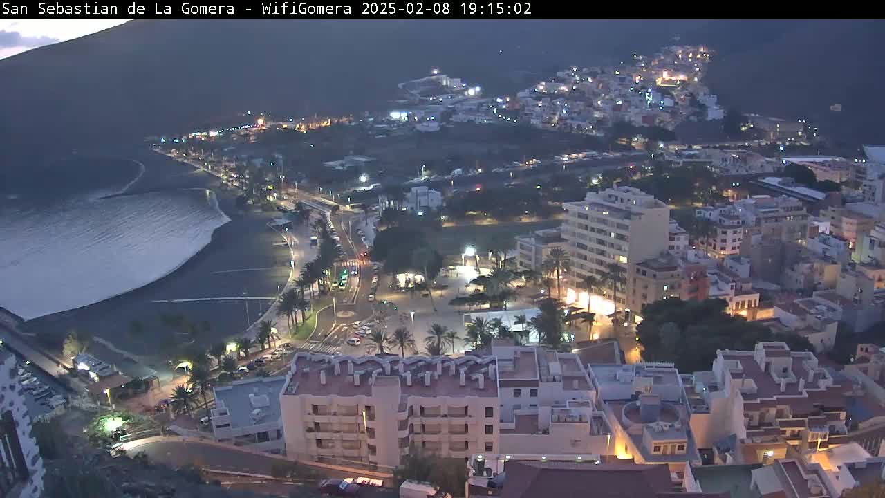A coastal town at night, with dark ocean waves lapping a black sand beach, under a mostly clear, dark sky.
