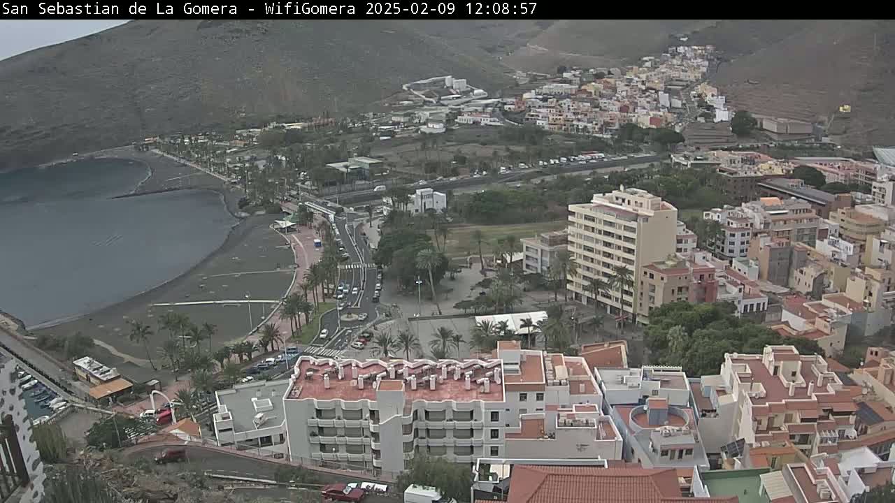 An overcast day reveals a coastal town nestled against a mountainous backdrop, featuring a dark-sand beach bordering a calm bay and numerous buildings of varying heights.