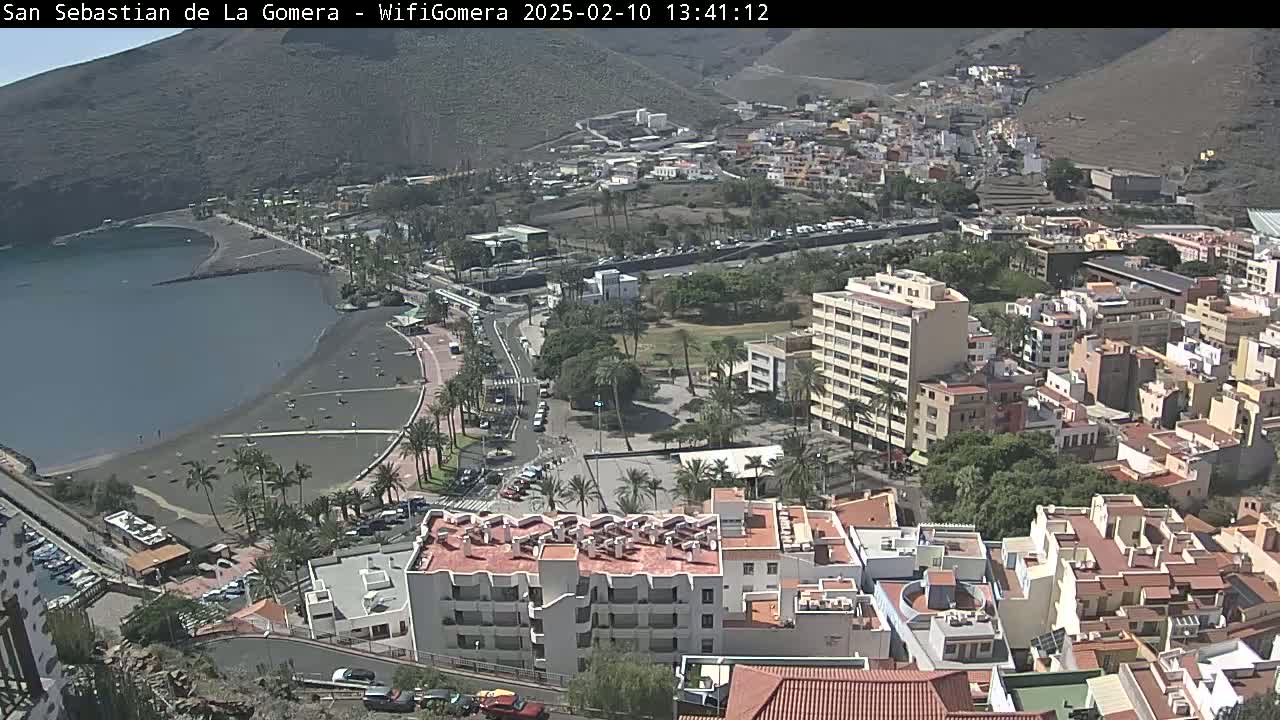 An elevated view shows a coastal town on a sunny day, with a dark-sand beach bordering a calm bay and buildings stretching up a hillside.
