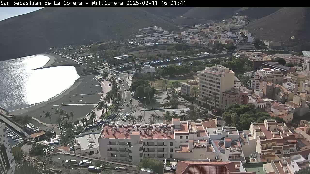 A coastal town with a dark-sand beach and calm ocean is nestled against a hillside, under a mostly sunny sky.