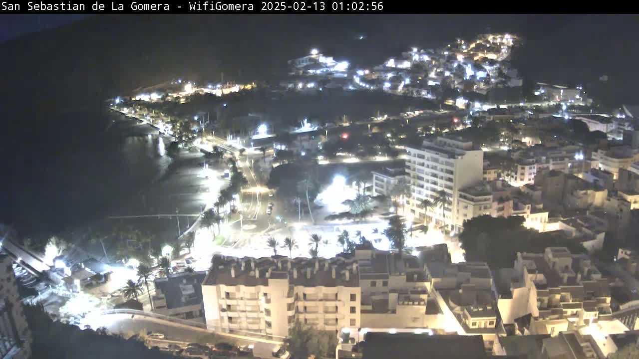 A nighttime aerial view of a coastal town, illuminated by streetlights, with a dark body of water visible in the foreground.