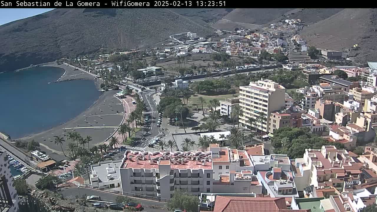 A coastal town with a dark-sand beach and calm ocean, nestled against a mountainside under sunny skies, is viewed from an elevated perspective.
