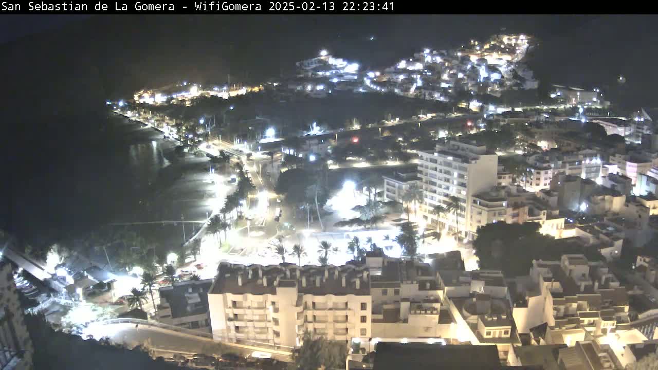 A nighttime aerial view of a coastal town shows brightly lit buildings clustered around a dark beach and calm ocean.