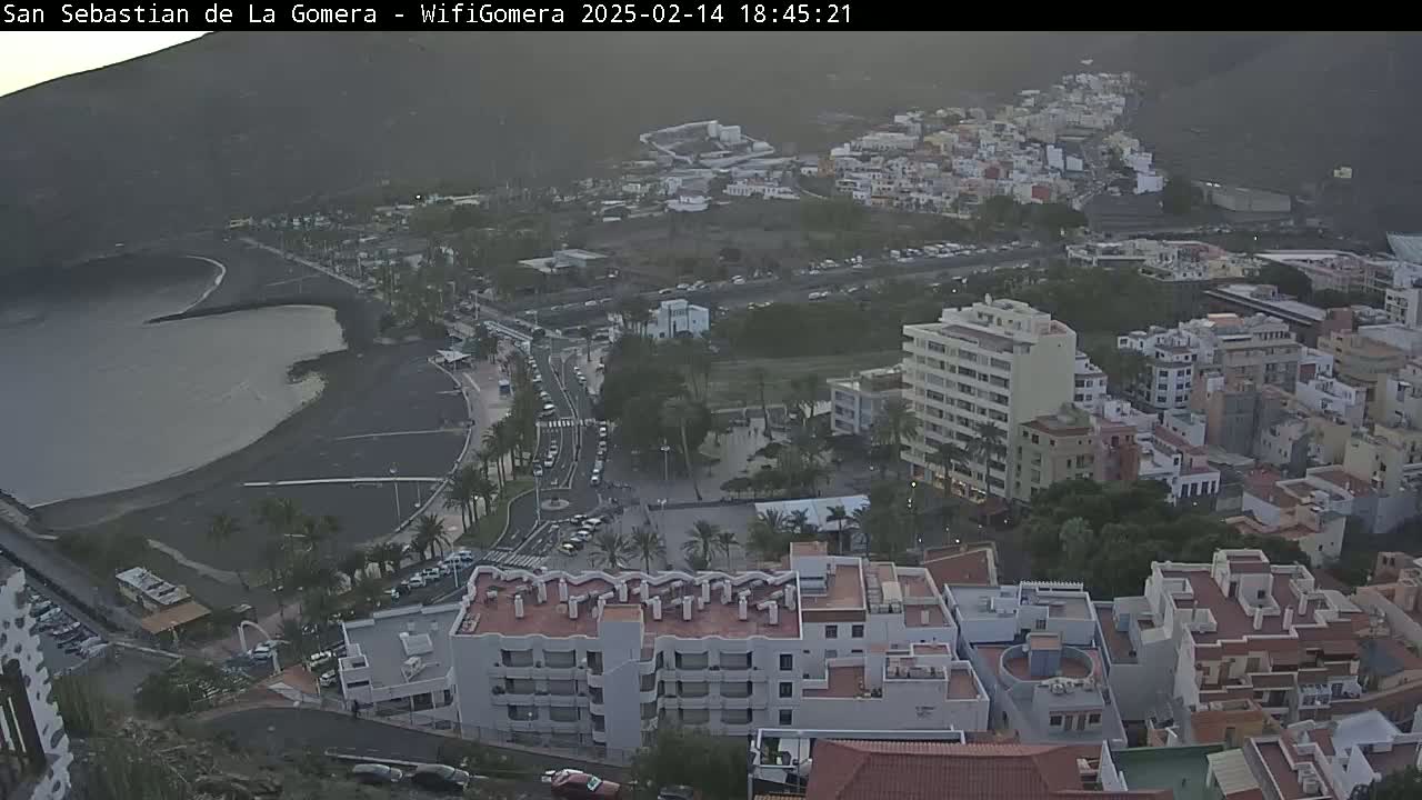An elevated view of a coastal town shows a dark-sand beach, buildings clustered together, and a calm body of water under an overcast sky.