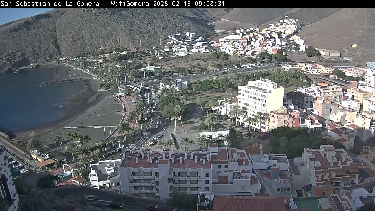 A coastal town with a dark-sand beach and numerous buildings is seen on a sunny day.