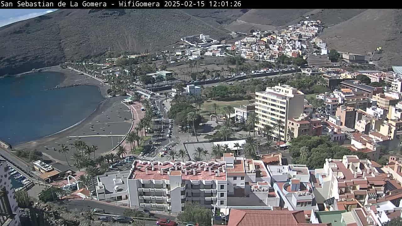 An aerial view shows a coastal town on a sunny day, with a dark-sand beach bordering a calm bay, and buildings extending uphill toward brown mountains.