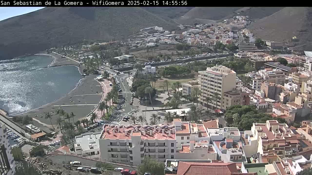 An aerial view shows a coastal town with a dark-sand beach and calm ocean under sunny skies, surrounded by buildings and palm trees.