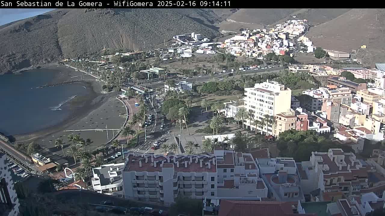 A coastal town on a sunny day, featuring a dark-sand beach, calm ocean waters, and buildings nestled against a hillside.
