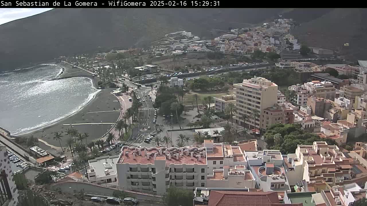 A coastal town with a dark sand beach and ocean waves, under a mostly sunny sky, is viewed from a high vantage point, showing buildings and streets extending into the hills.