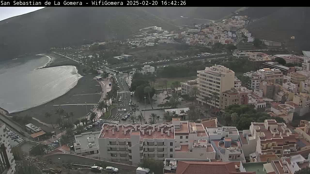 An overcast coastal town with a dark-sand beach and moderate wave action is viewed from a high vantage point.