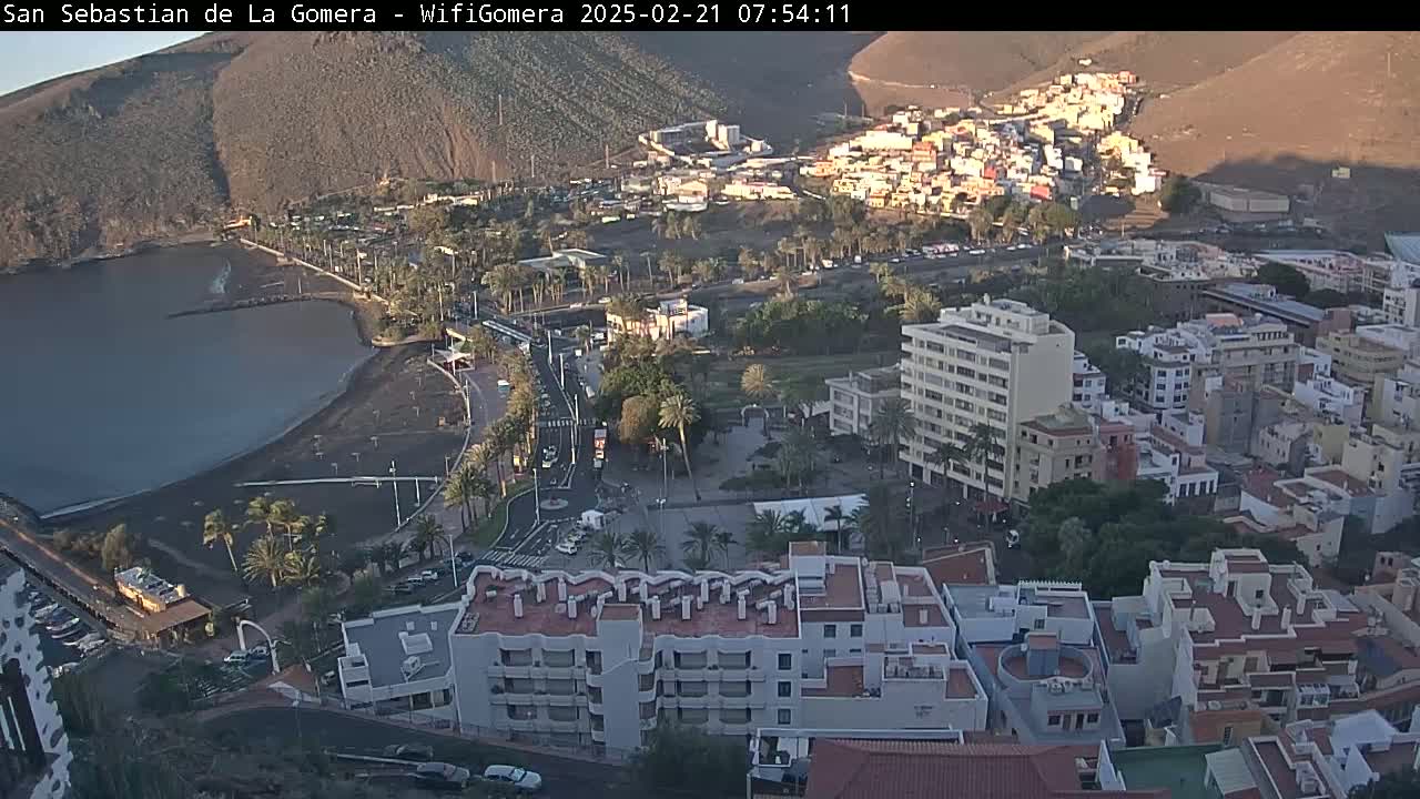 An aerial view of a coastal town at sunrise, showing a dark-sand beach, buildings, palm trees, and calm water.