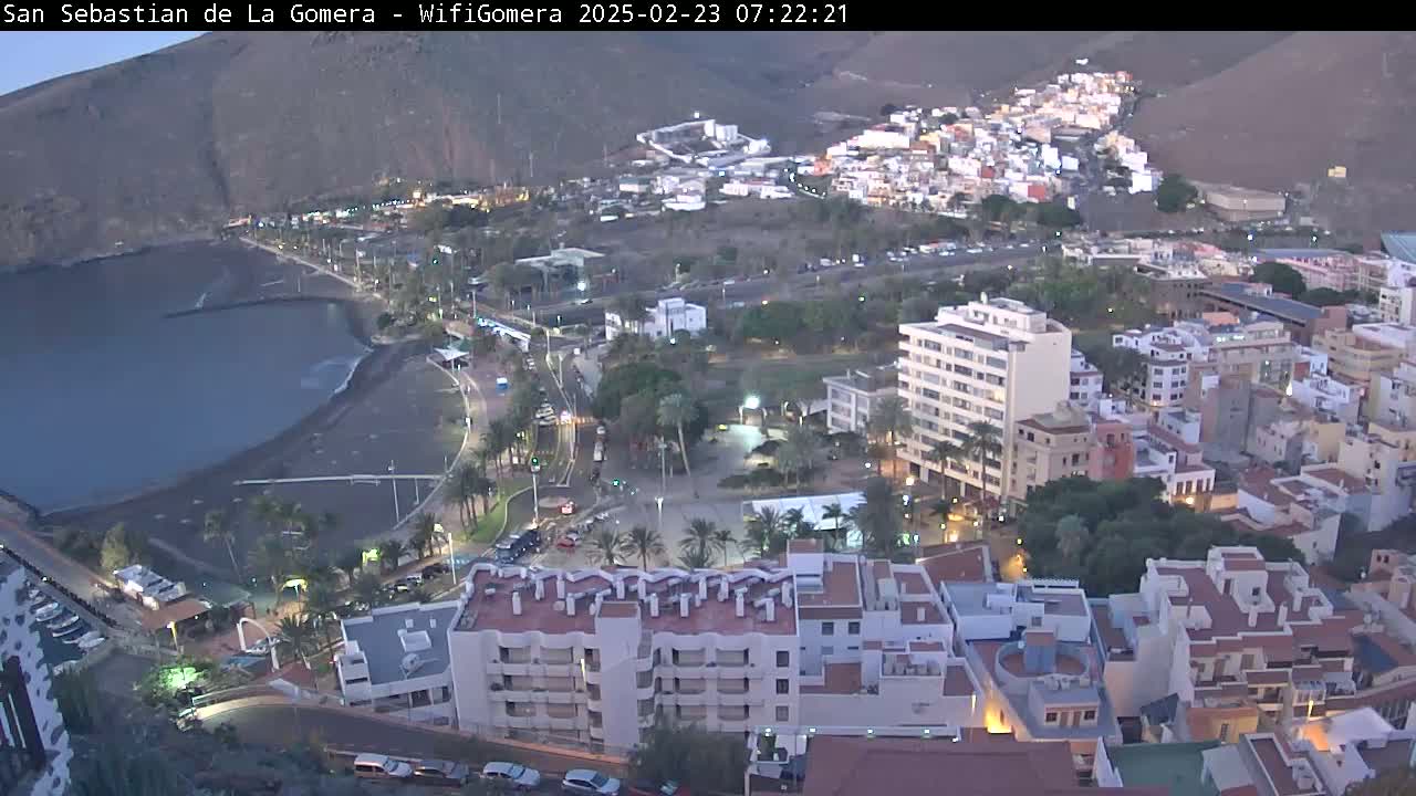 An aerial evening view of a coastal town with dark-colored sand beach, buildings, and palm trees, under a twilight sky.