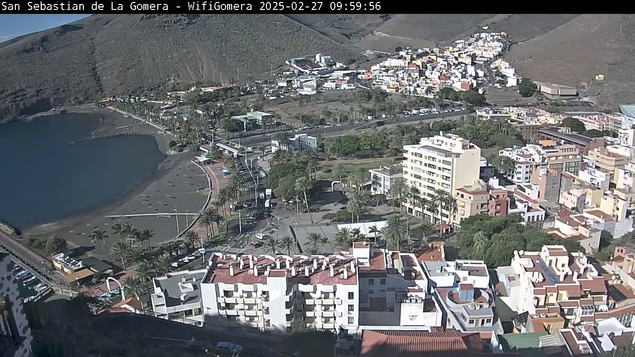 An aerial view shows a coastal town on a sunny day, featuring a dark-sand beach, a calm bay, and numerous buildings clustered around a central plaza.