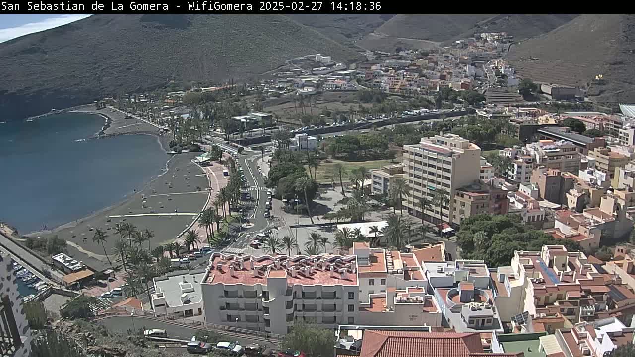 An aerial view of a coastal town on a sunny day, showing a dark-sand beach, calm bay, buildings, roads, and palm trees.