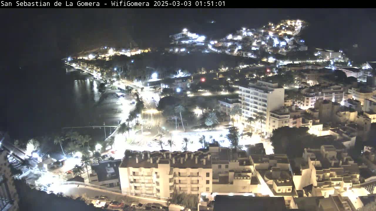 A nighttime aerial view of a coastal town with buildings illuminated, streets showing vehicle lights, and a dark body of water visible in the background.