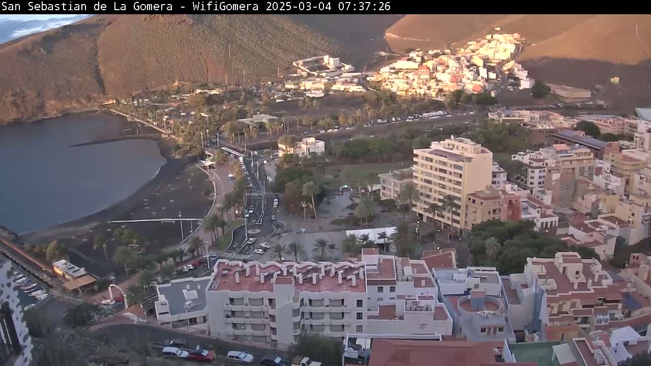 An aerial view shows a coastal town at sunrise, featuring a dark-sand beach, calm water, and buildings of varying heights, all set against a backdrop of sunlit hills.