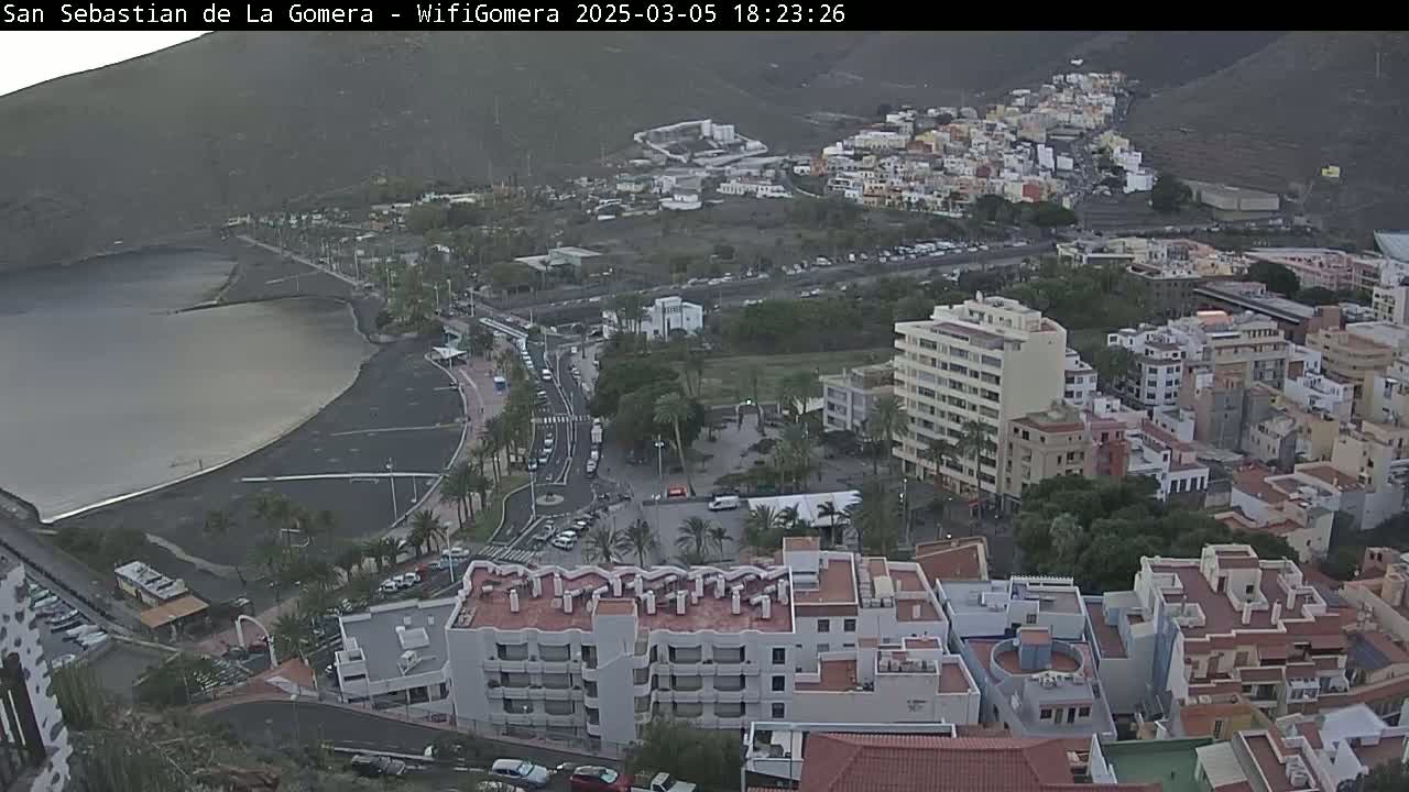 An aerial view shows a coastal town with buildings clustered around a dark-sand beach and calm bay under an overcast sky.