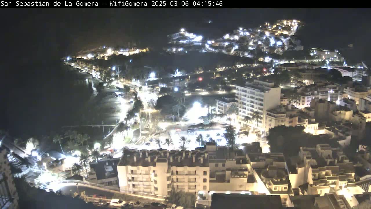 A nighttime aerial view of a coastal town, illuminated with streetlights, showing buildings, roads, and a dark body of water along the shoreline.