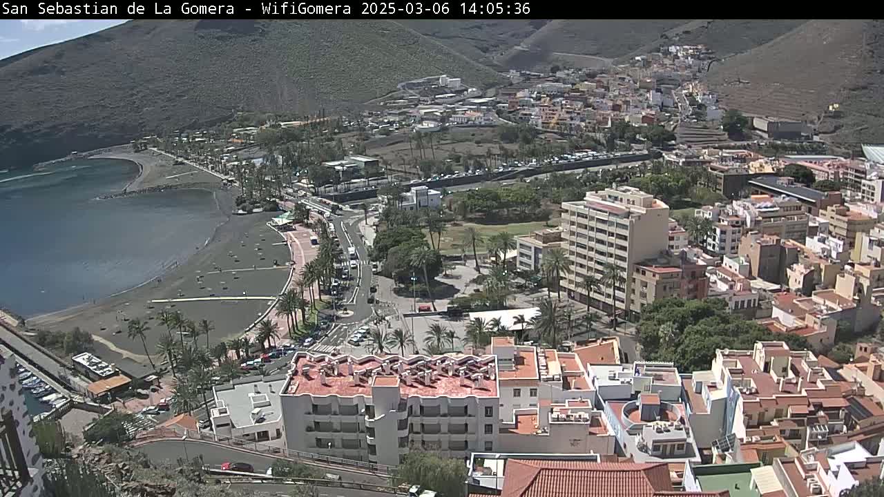 A coastal town with a black sand beach and numerous buildings stretches along a hillside under sunny skies.