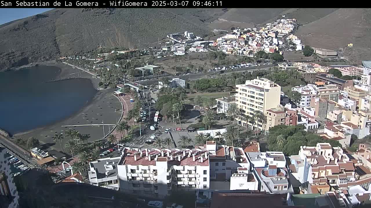 An aerial view of a coastal town on a sunny day shows a dark-sand beach, a calm body of water, and numerous buildings, including apartment complexes and hotels, nestled against a hillside.