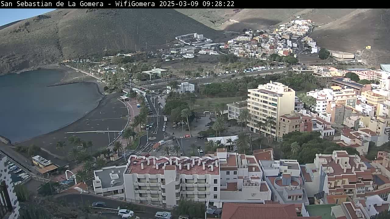 An aerial view shows a coastal town nestled against a volcanic hillside on a partly sunny day, with a dark-colored beach bordering a calm body of water.