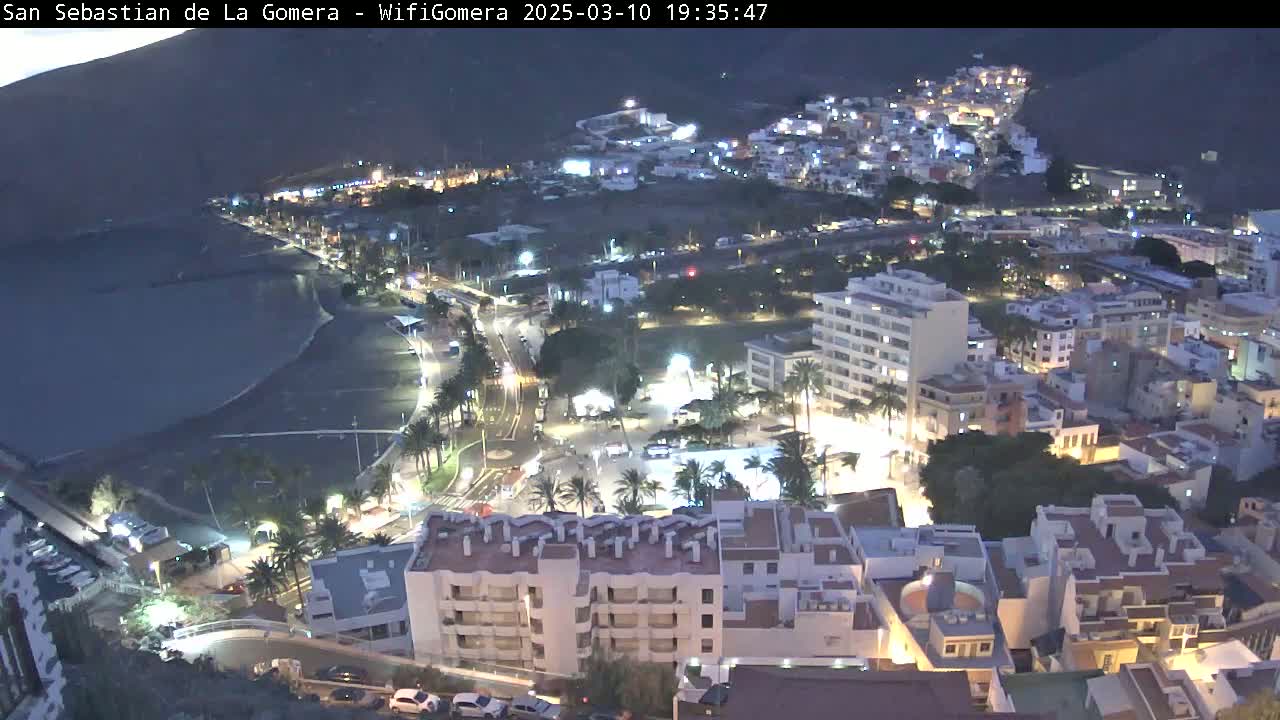 A nighttime aerial view of a coastal town, showing buildings lit up along a dark ocean shore under a clear, dark sky.
