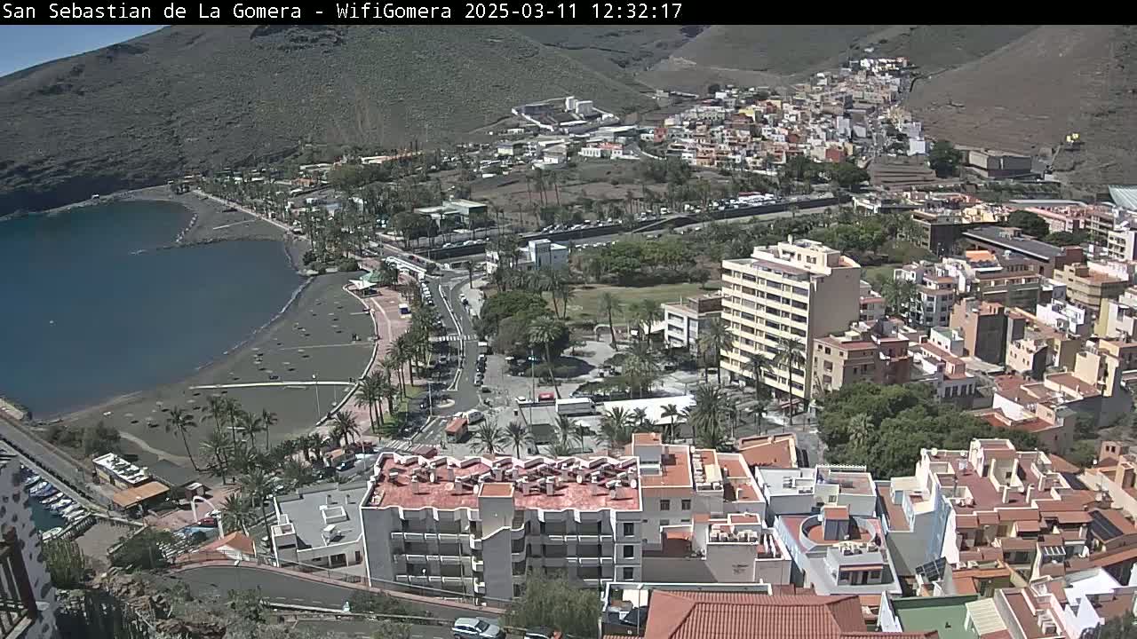 A coastal town with a dark-sand beach and calm ocean under sunny skies is viewed from a high vantage point, showing many buildings nestled against a hillside.