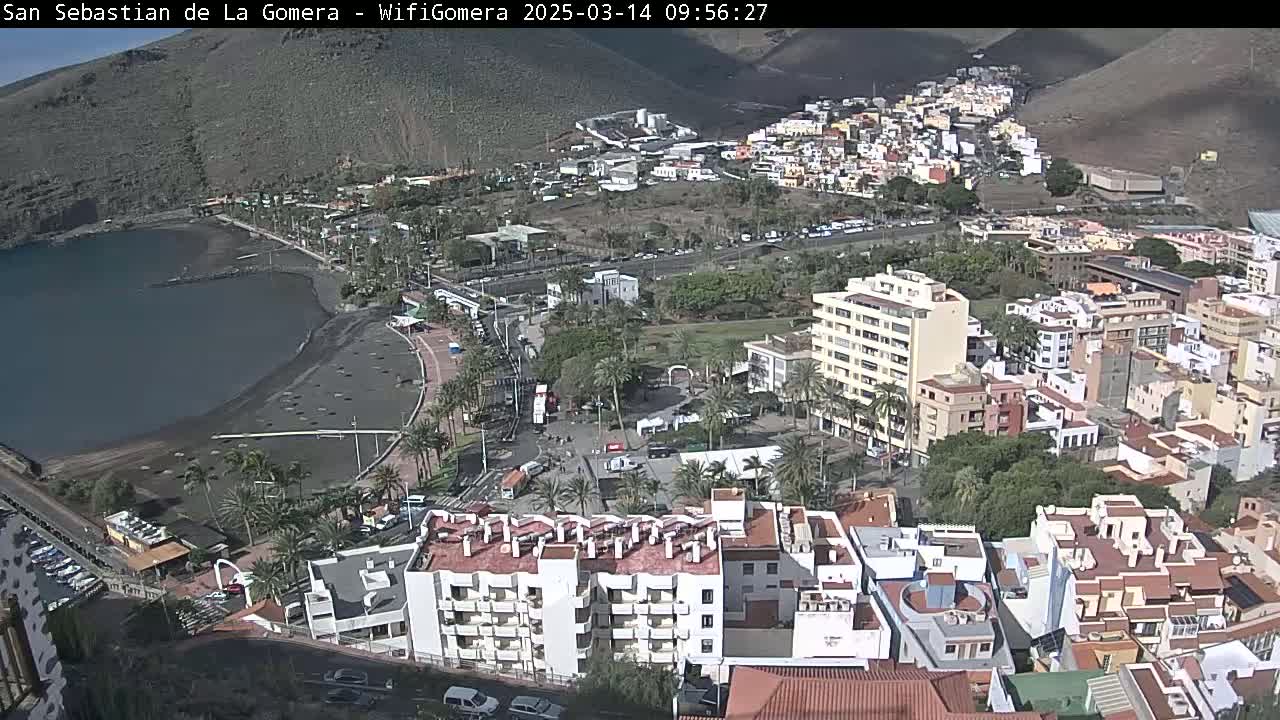 An aerial view of a coastal town on a sunny day, showing a black sand beach, a calm bay, and numerous buildings nestled against a hillside.