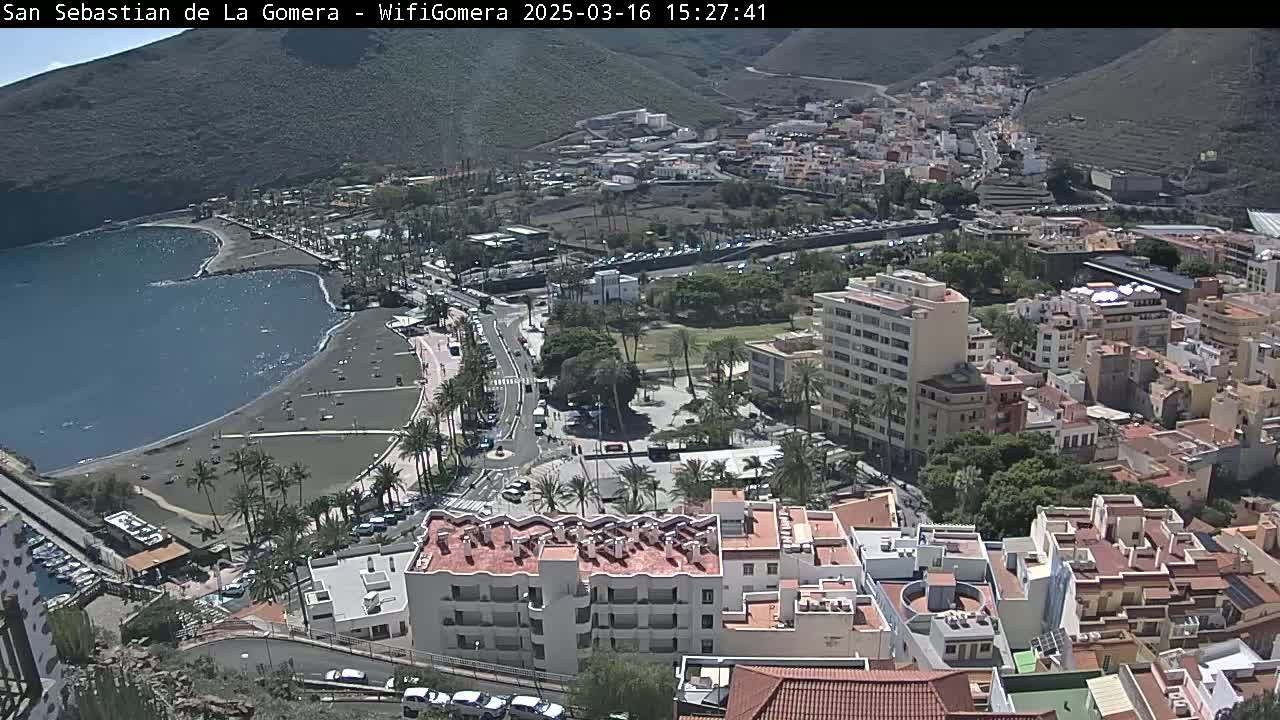 A coastal town with a dark-sand beach and calm waters nestled against a mountainside on a sunny day.