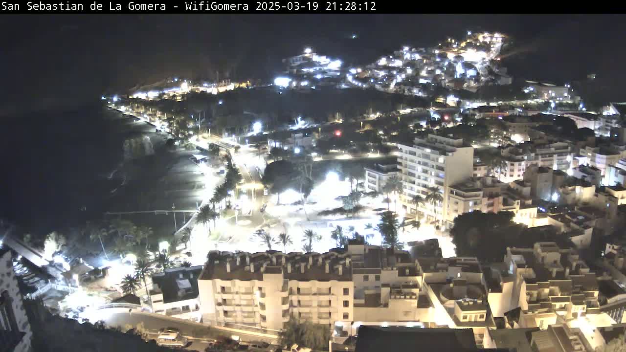 A nighttime, aerial view of a coastal town with buildings illuminated, a dark ocean, and a beach.