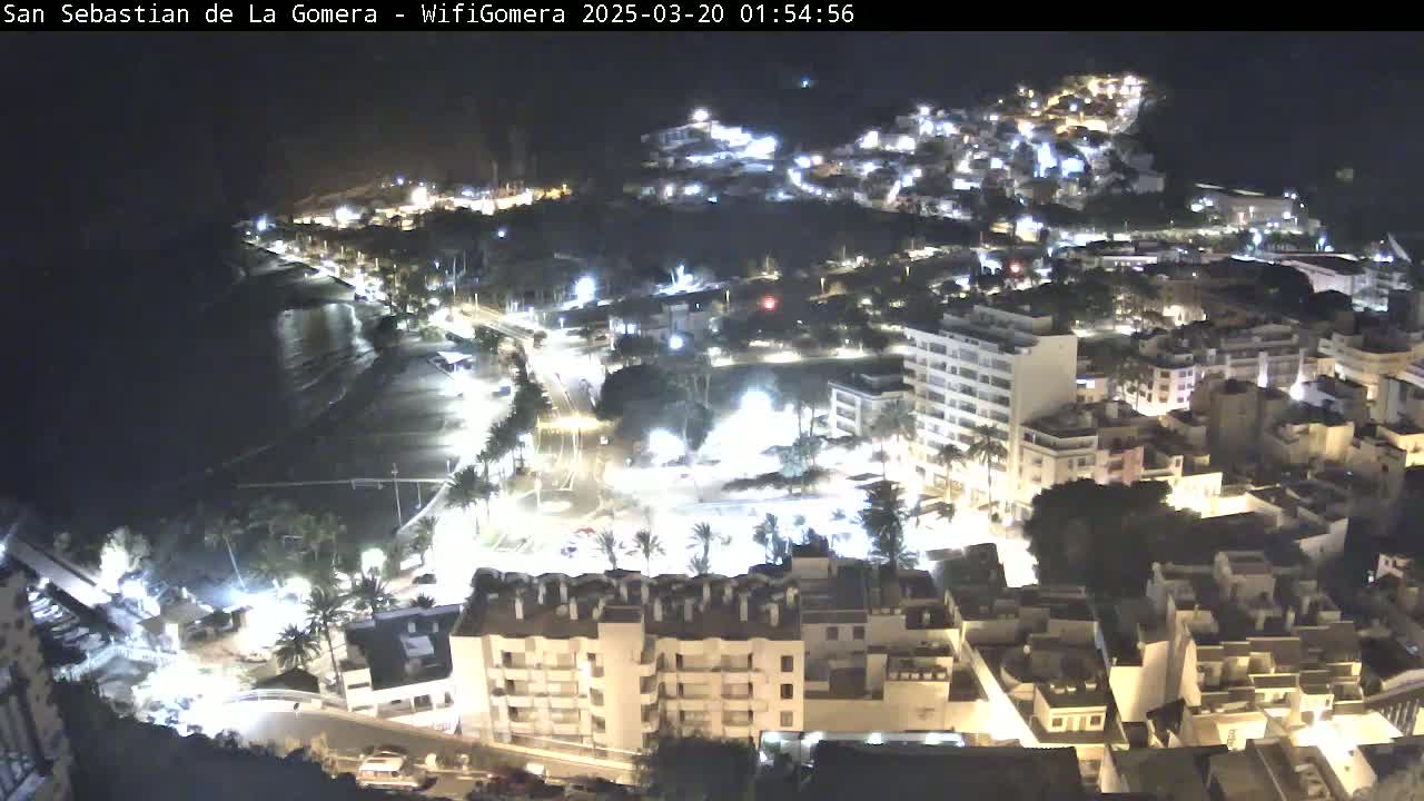 A nighttime aerial view of a coastal town with brightly lit buildings, streets, and a dark beach.