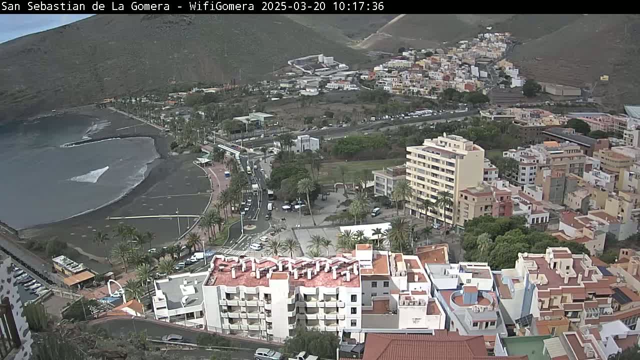 An aerial view of a coastal town on a partly sunny day shows a black sand beach with small waves, buildings clustered together, and a road winding along the waterfront.
