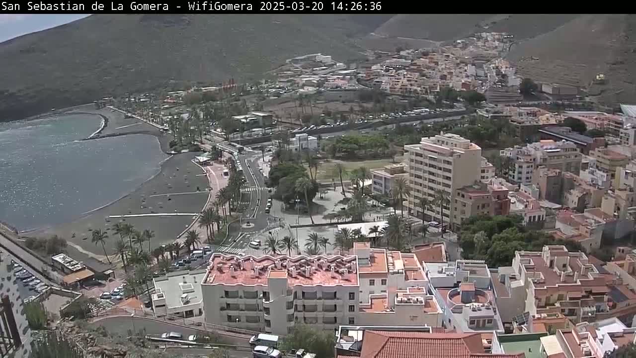 An aerial view shows a coastal town on a sunny day, with a dark-sand beach bordering a calm body of water and buildings rising up a hillside.