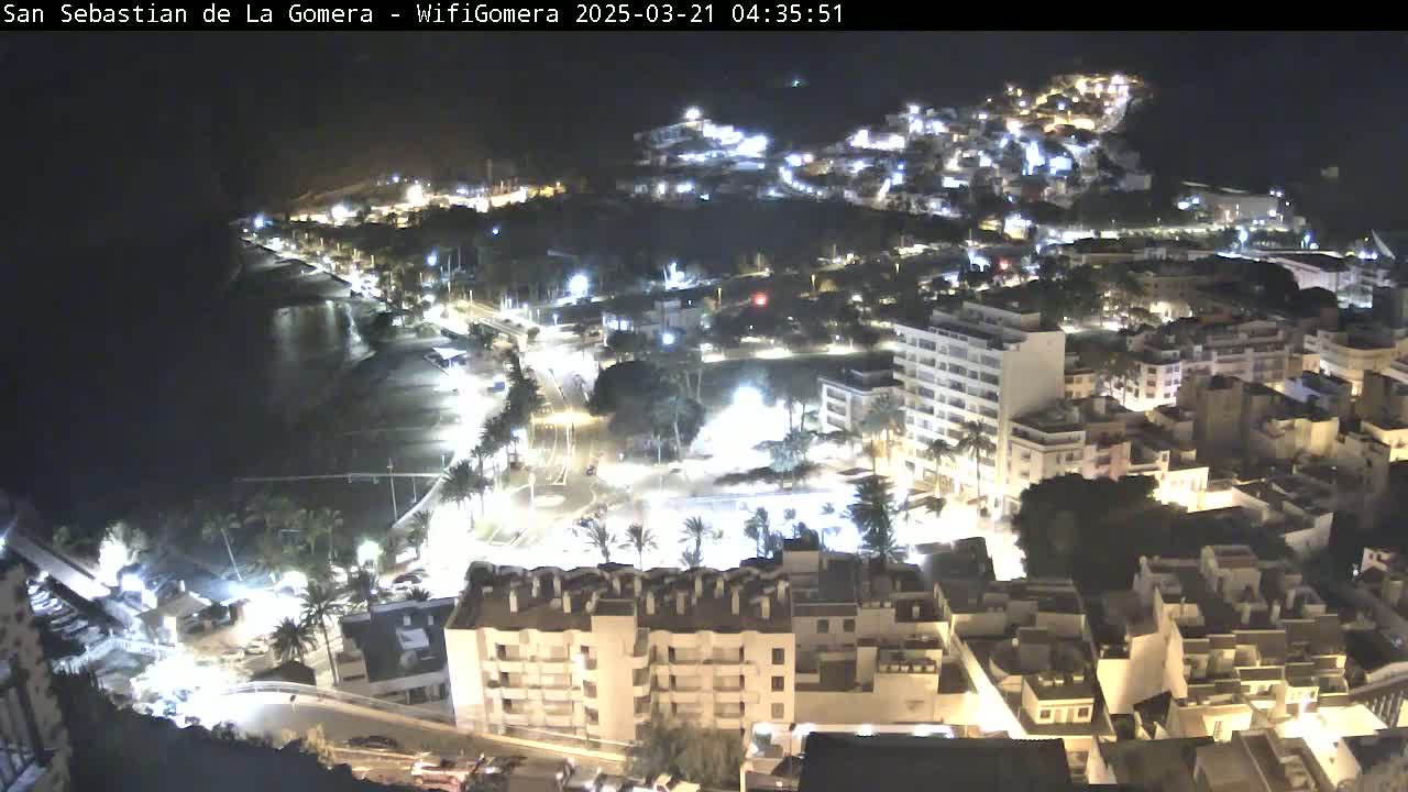 A nighttime aerial view of a coastal town, brightly lit buildings sprawling across a hillside overlooking a dark beach and ocean.