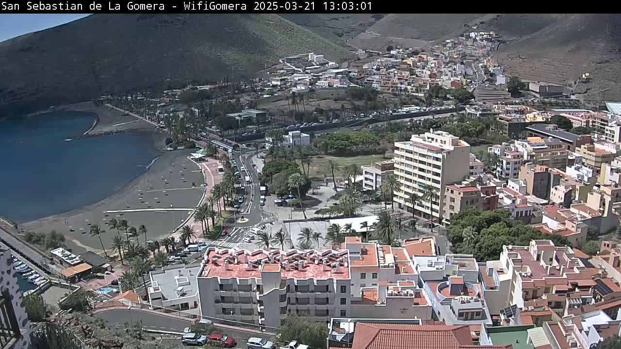 A coastal town with a black sand beach and numerous buildings stretches along a bay under a sunny sky.
