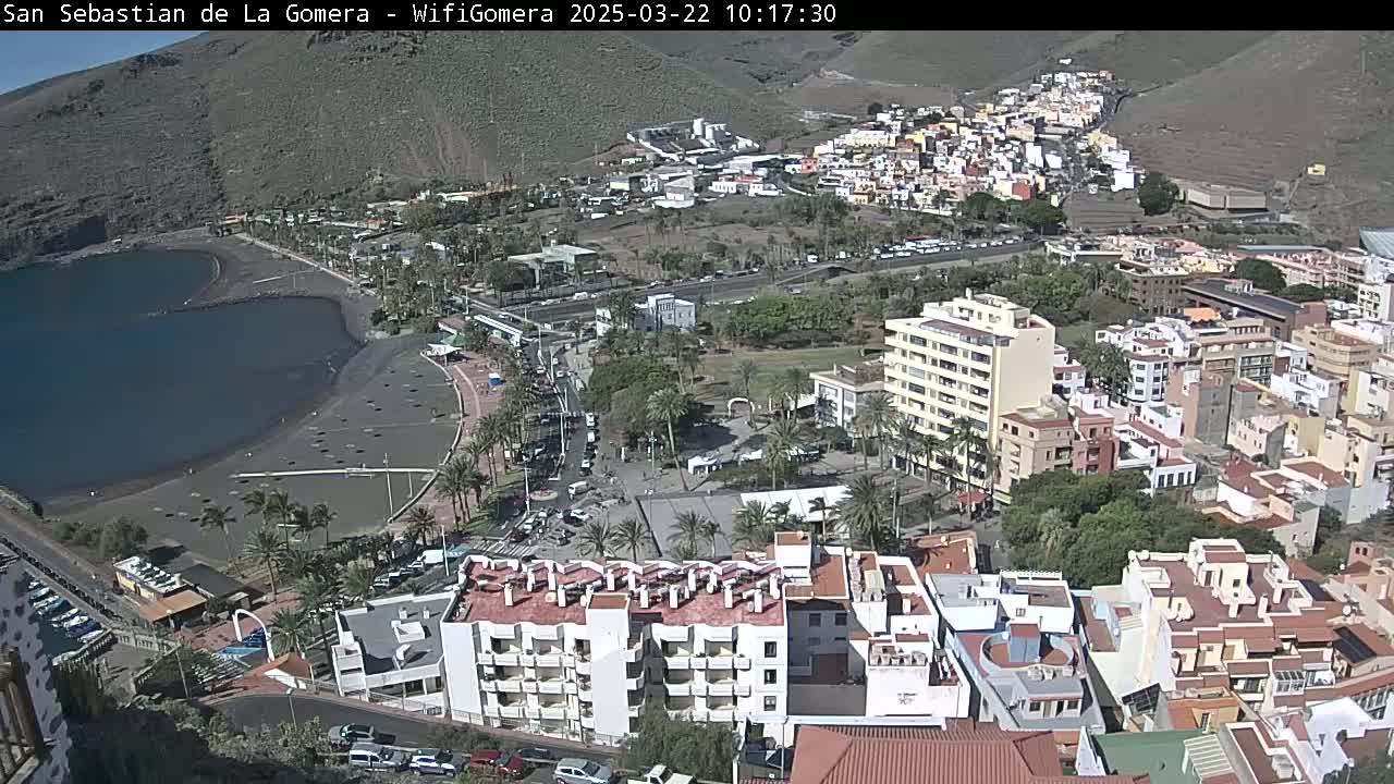 An aerial view shows a coastal town on a sunny day, featuring a dark-sand beach bordering a calm body of water, and numerous buildings stretching up a hillside.