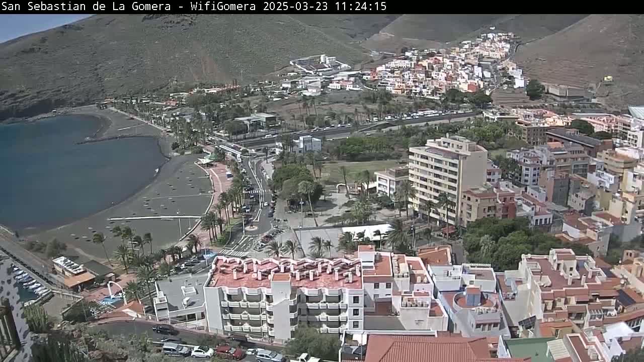 A coastal town with dark sand beach and buildings, situated on a hillside under sunny skies.