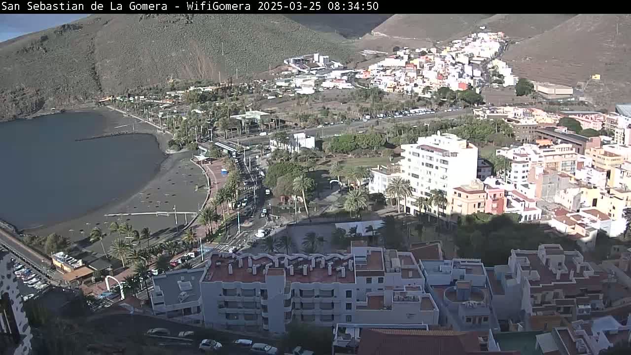 A coastal town nestled against a mountainside under sunny skies shows a bay, beach, buildings, palm trees, and roads.