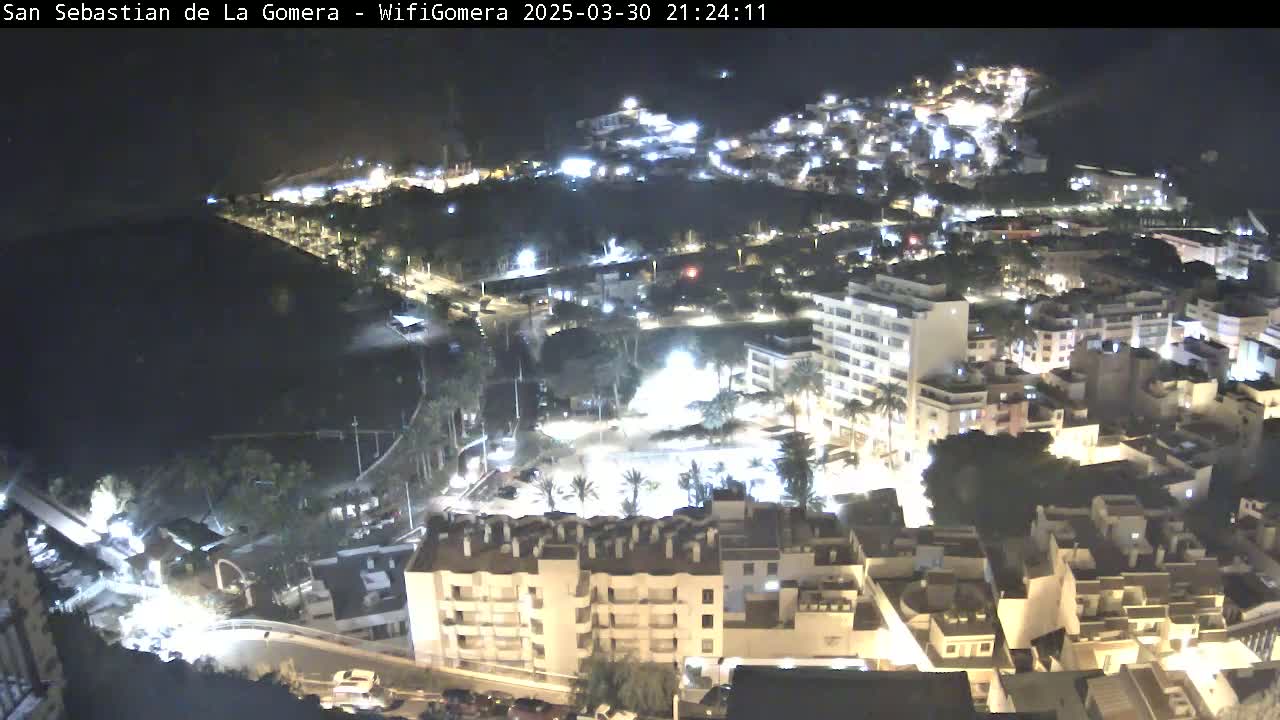 A nighttime aerial view of a coastal town shows many buildings lit up, with a dark body of water visible in the background.