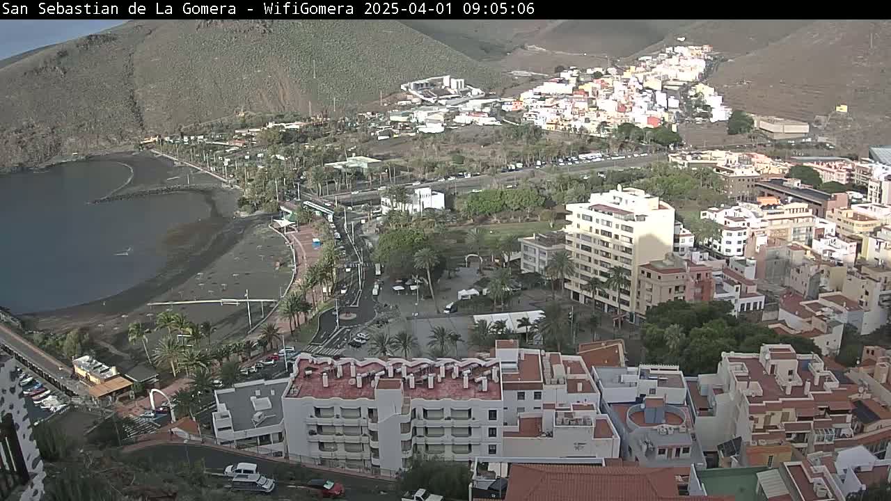 A coastal town with a black sand beach and numerous buildings is visible under a mostly clear sky.