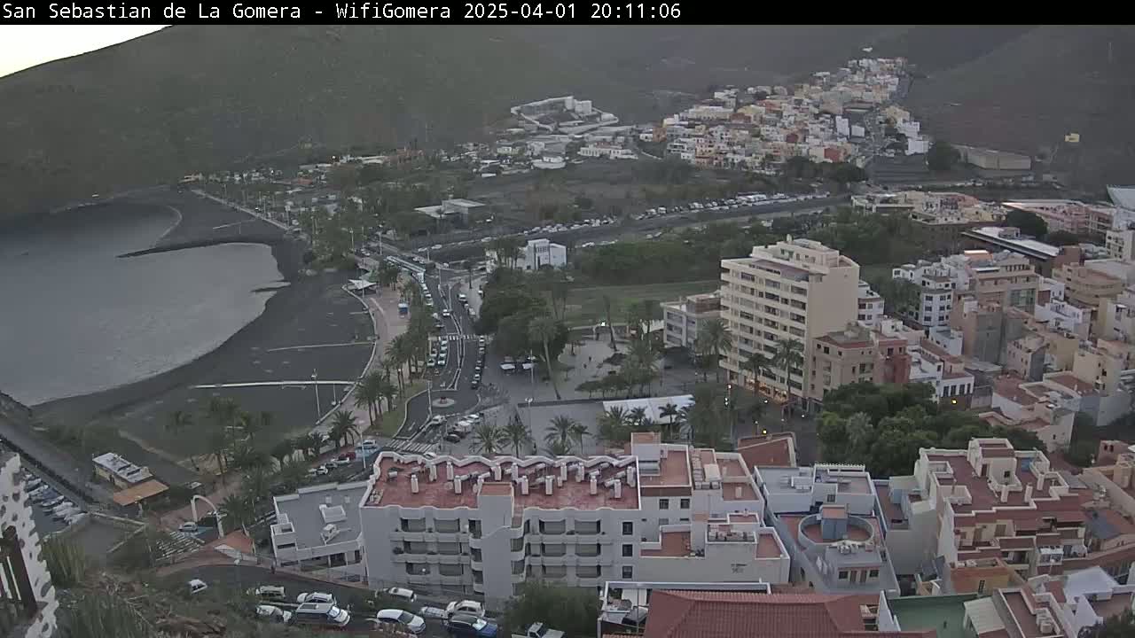 An aerial view shows a coastal town at dusk, with a dark-sand beach curving into a calm bay,  surrounded by low-rise buildings and palm trees.