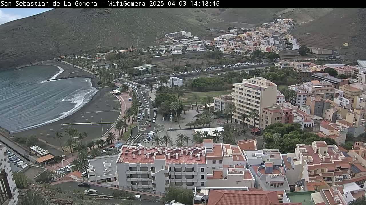 An overcast day reveals a coastal town nestled against a hillside, featuring a dark-sand beach with gentle waves and a collection of buildings ranging from low-rise structures to taller apartment complexes.
